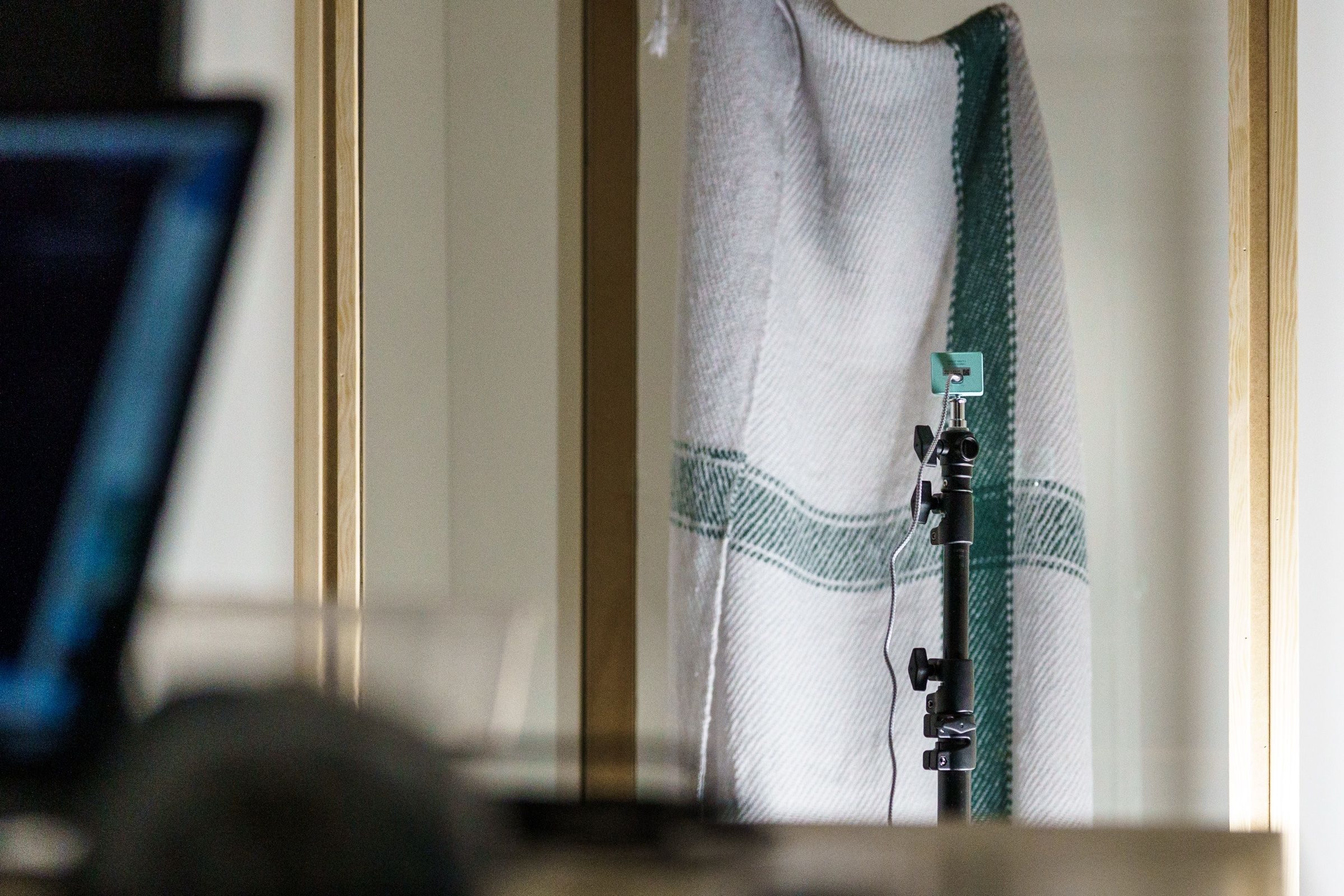 A small camera on a black stand in front of a glass cabinet, where a cream plaid with a green stripe hangs, a laptop blurred in the foreground