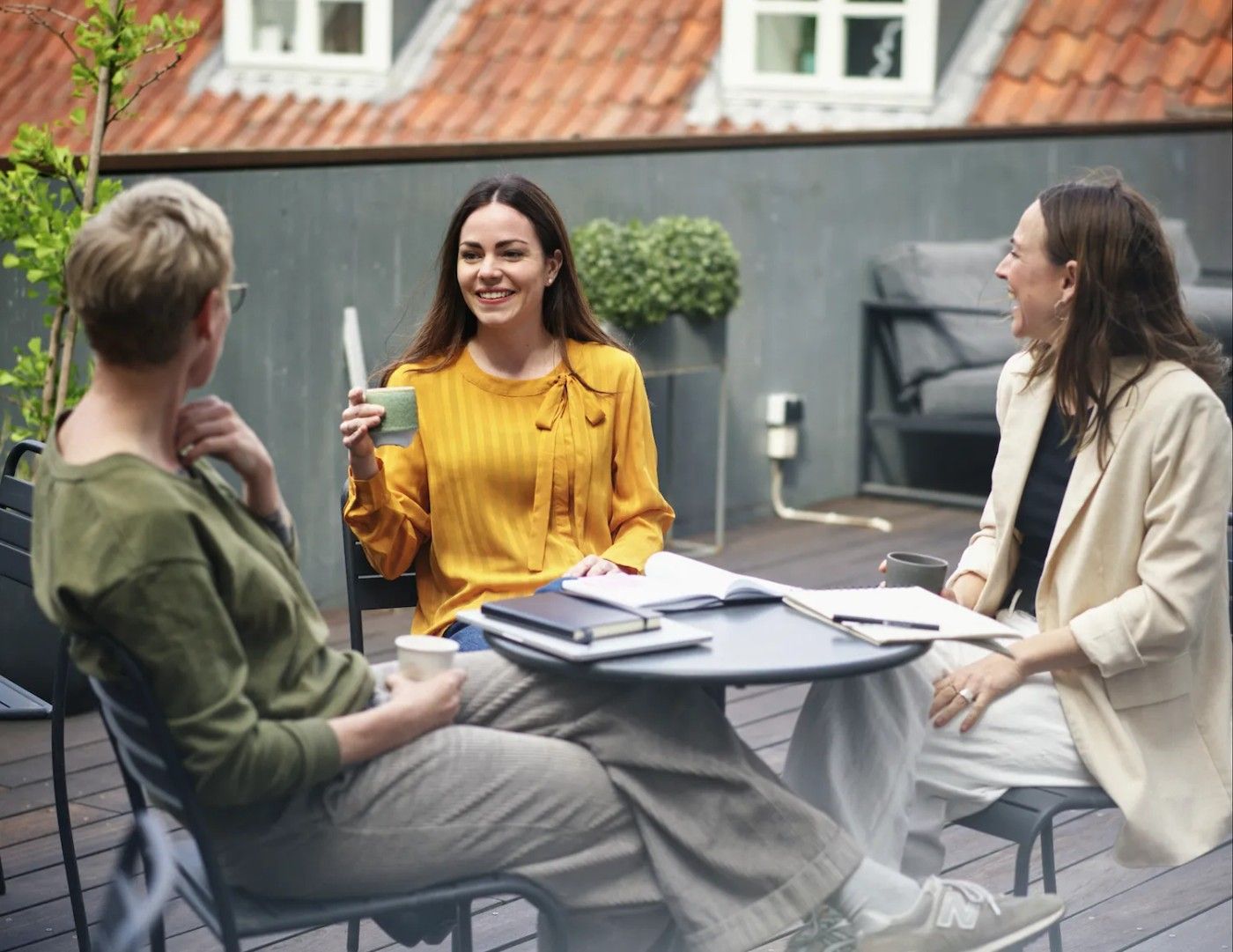 Three colleagues talking over coffee and notebooks at a table on a roof terrace, one laughing in a yellow jumper