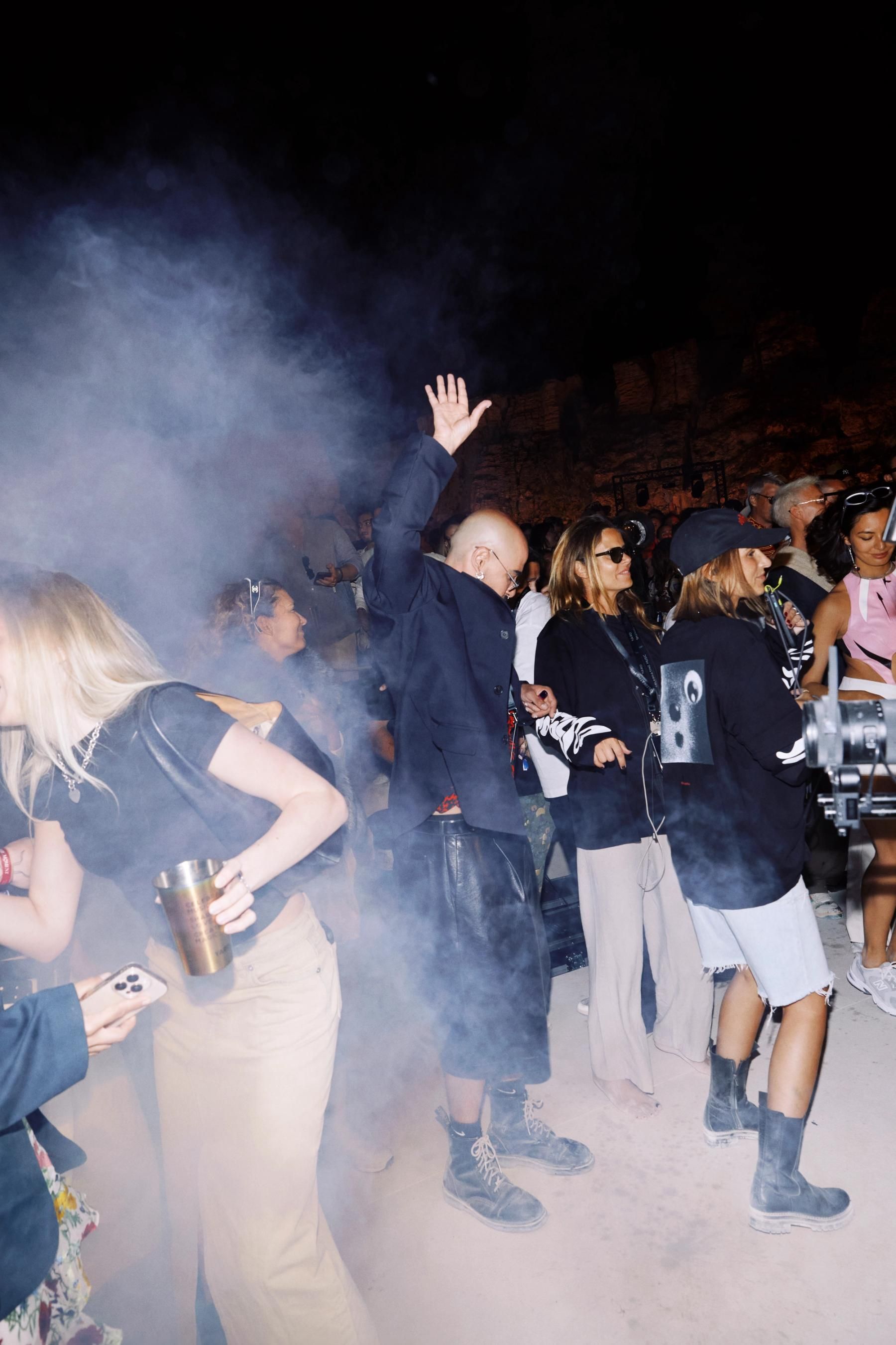 A crowd dancing in thick smoke at night under a floodlit stone wall, one man in a dark blazer with his hand raised high