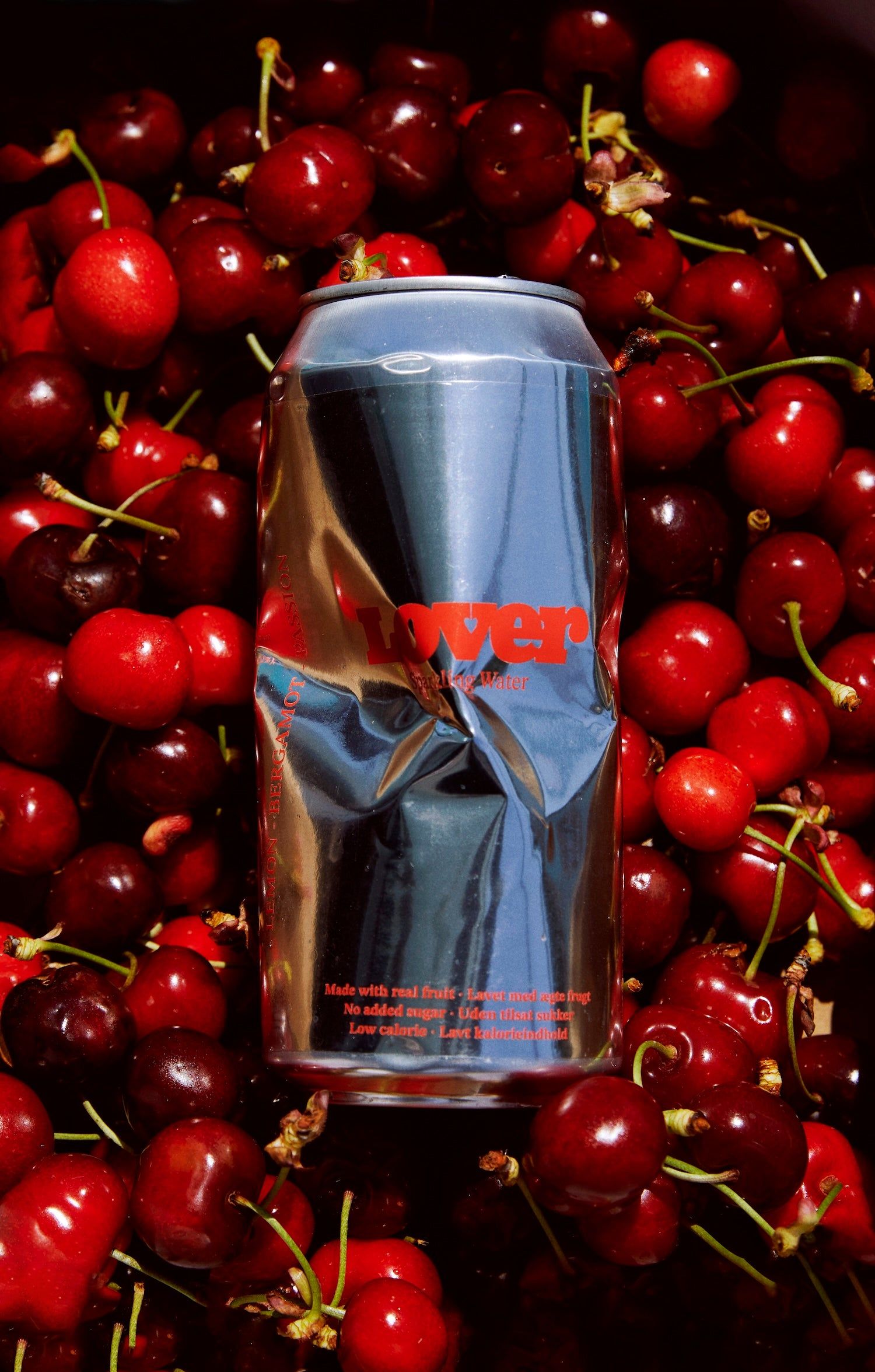 A crushed chrome Lover sparkling water can lying on a pile of glossy red cherries