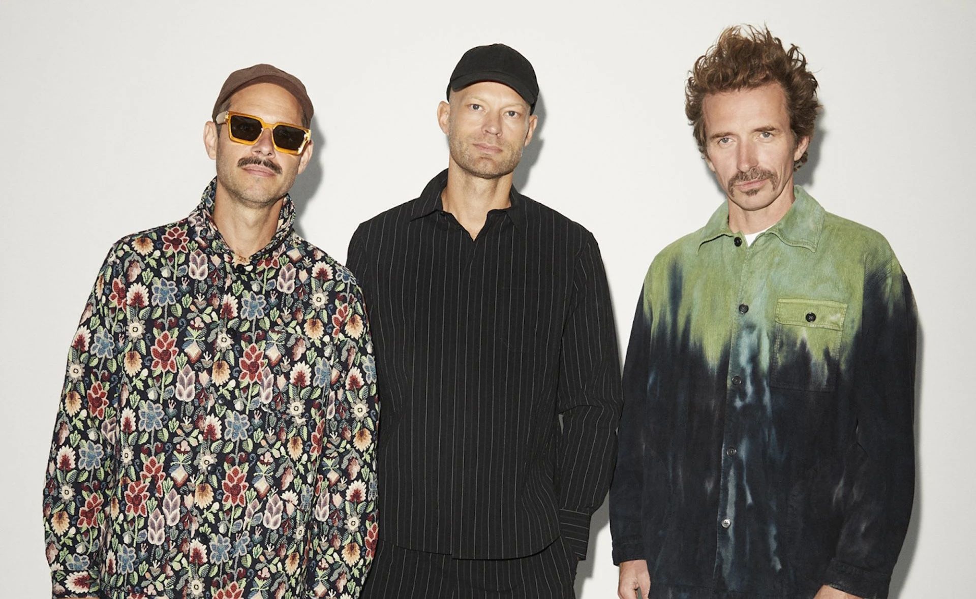 The three members of WhoMadeWho against a white wall, one in a floral jacket and orange sunglasses, one in black pinstripes and a cap, one in a green and black tie-dyed shirt