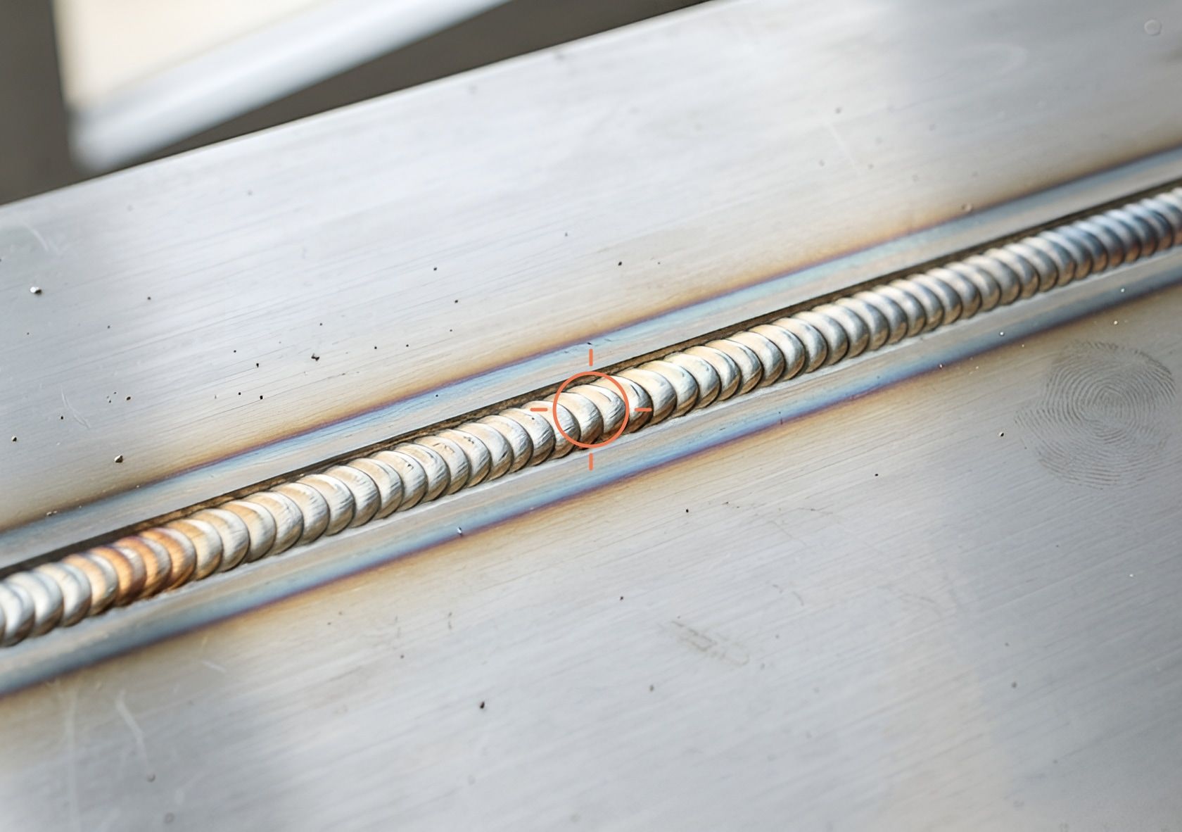 A close-up of a TIG weld seam on a brushed steel plate, one point on the bead circled by an orange detection marker