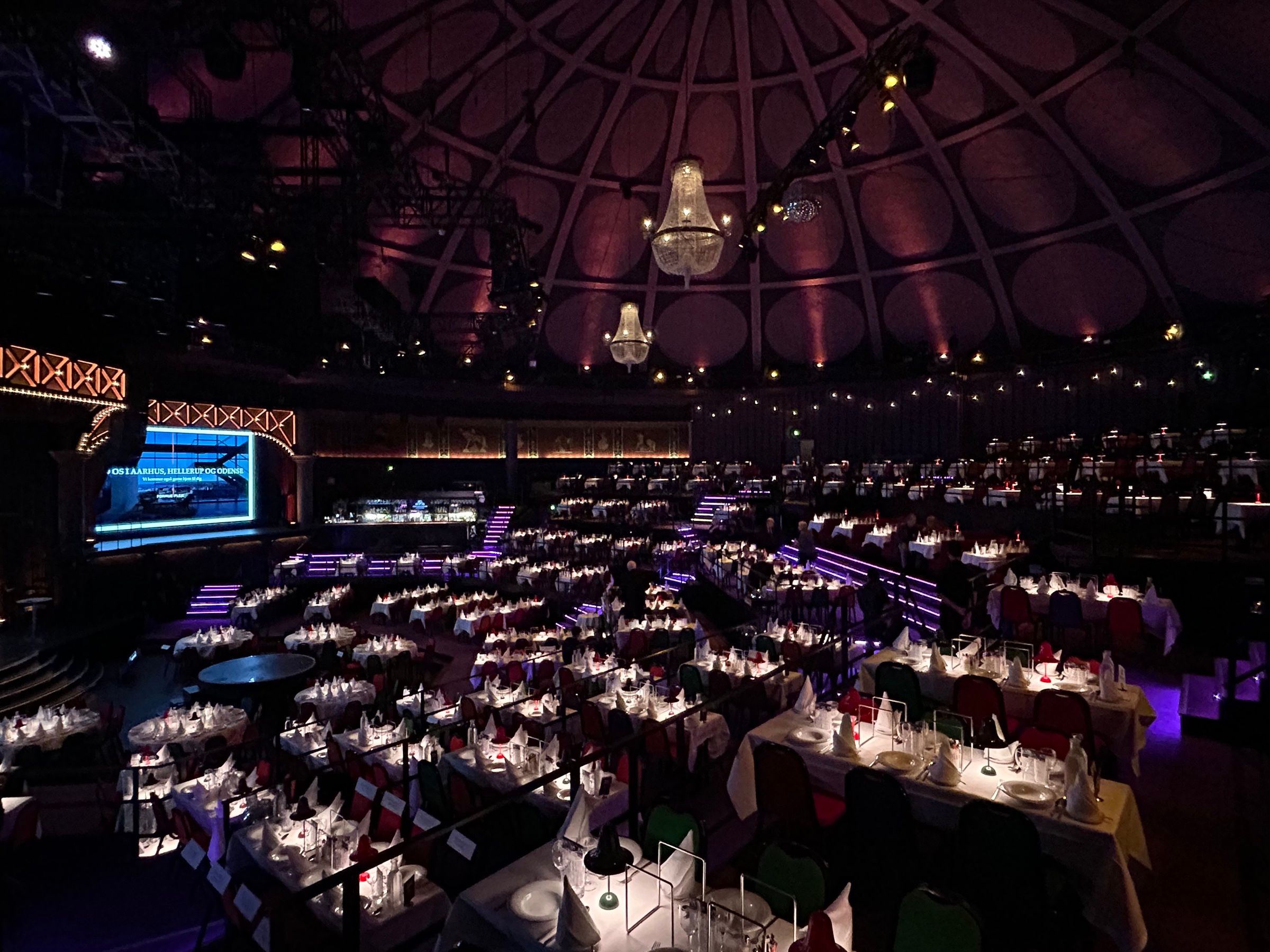 A dark dinner theatre under a painted dome, rows of white-clothed tables lit by chandeliers and a glowing stage screen